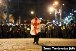 Протестующий, завернутый в флаг Грузии, идет навстречу полицейским, выстроившимся в шеренгу у здания парламента Грузии, Тбилиси, 7 марта 2023 года.